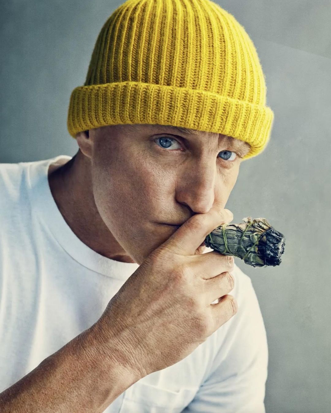 Close-up of adult male in yellow beanie holding a smoking sage bundle near his face against a neutral studio backdrop.