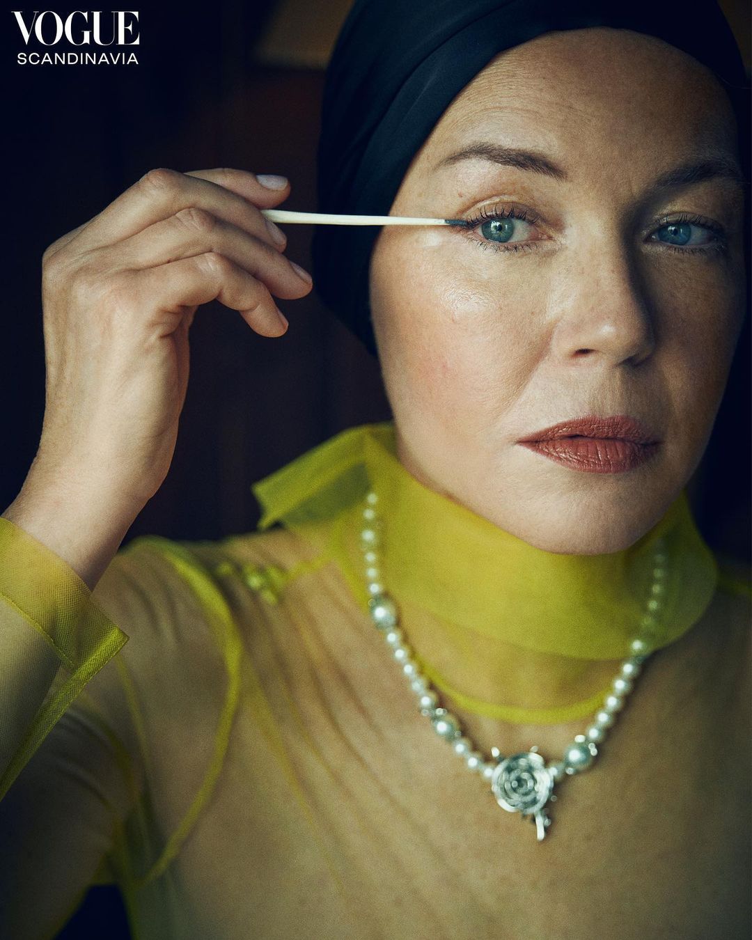 Close-up portrait of a stylish woman applying eye makeup, wearing a black head covering, pearl necklace, and yellow sheer top indoors.