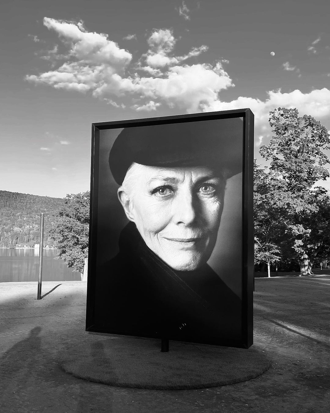 Black and white portrait of a woman in a framed photograph, displayed outdoors in a park setting with trees and lake visible.