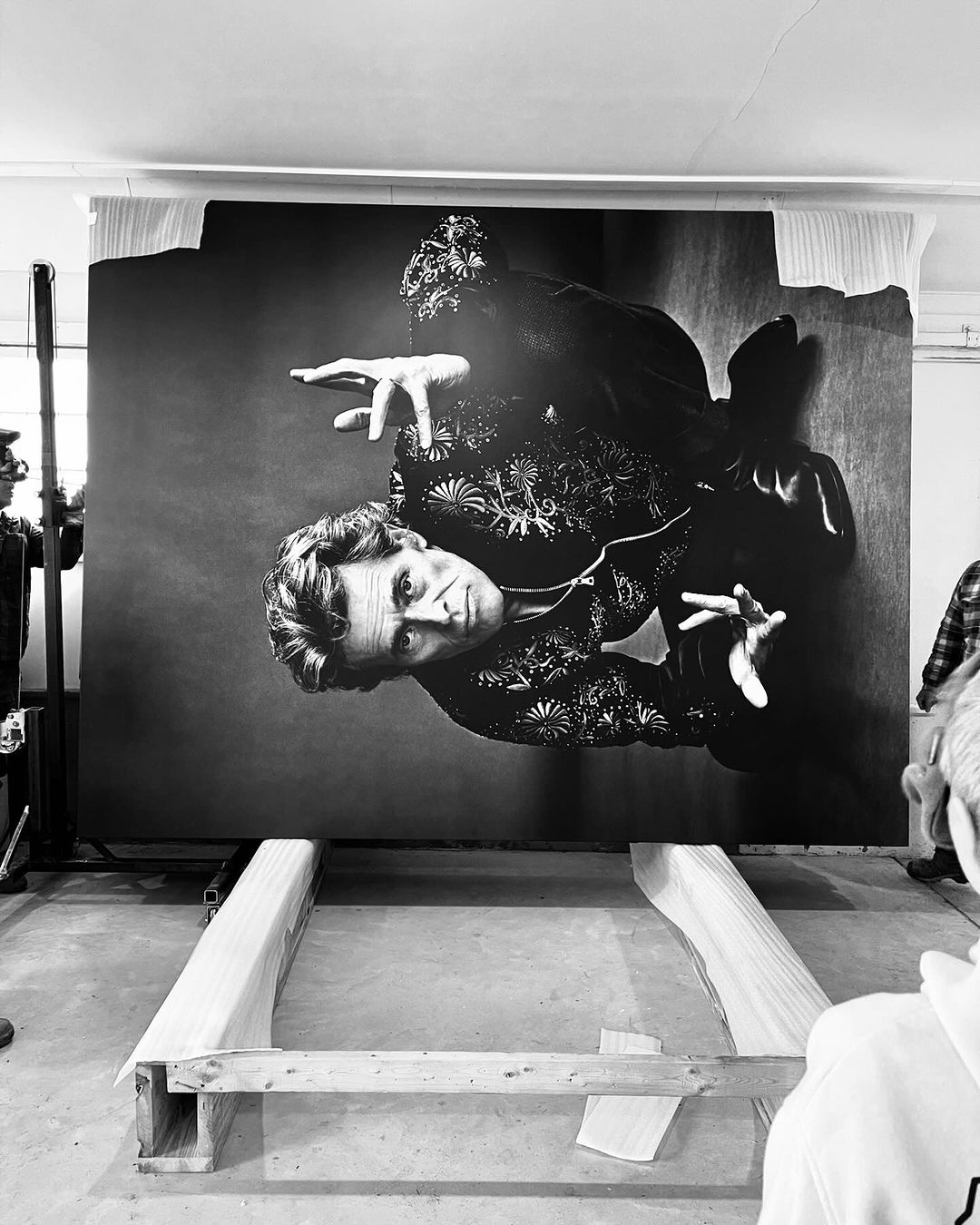 Black-and-white portrait of a man in an embroidered shirt on a large print propped on a wooden frame inside a white room.