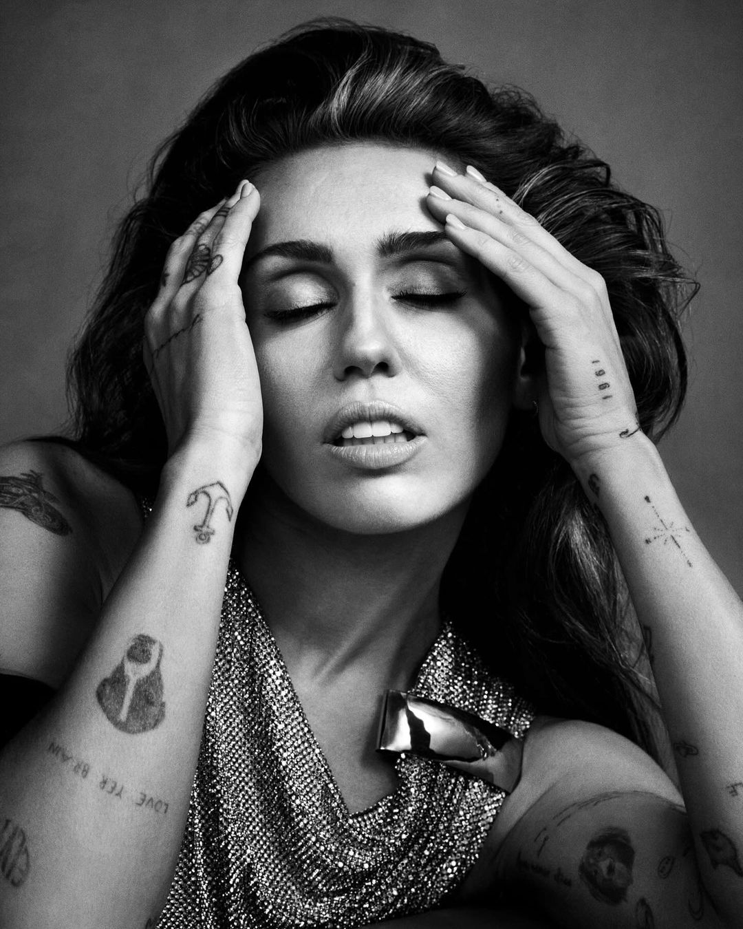 Black and white portrait of a woman with hands on her head and eyes closed, wearing a metallic top with a large, reflective brooch.