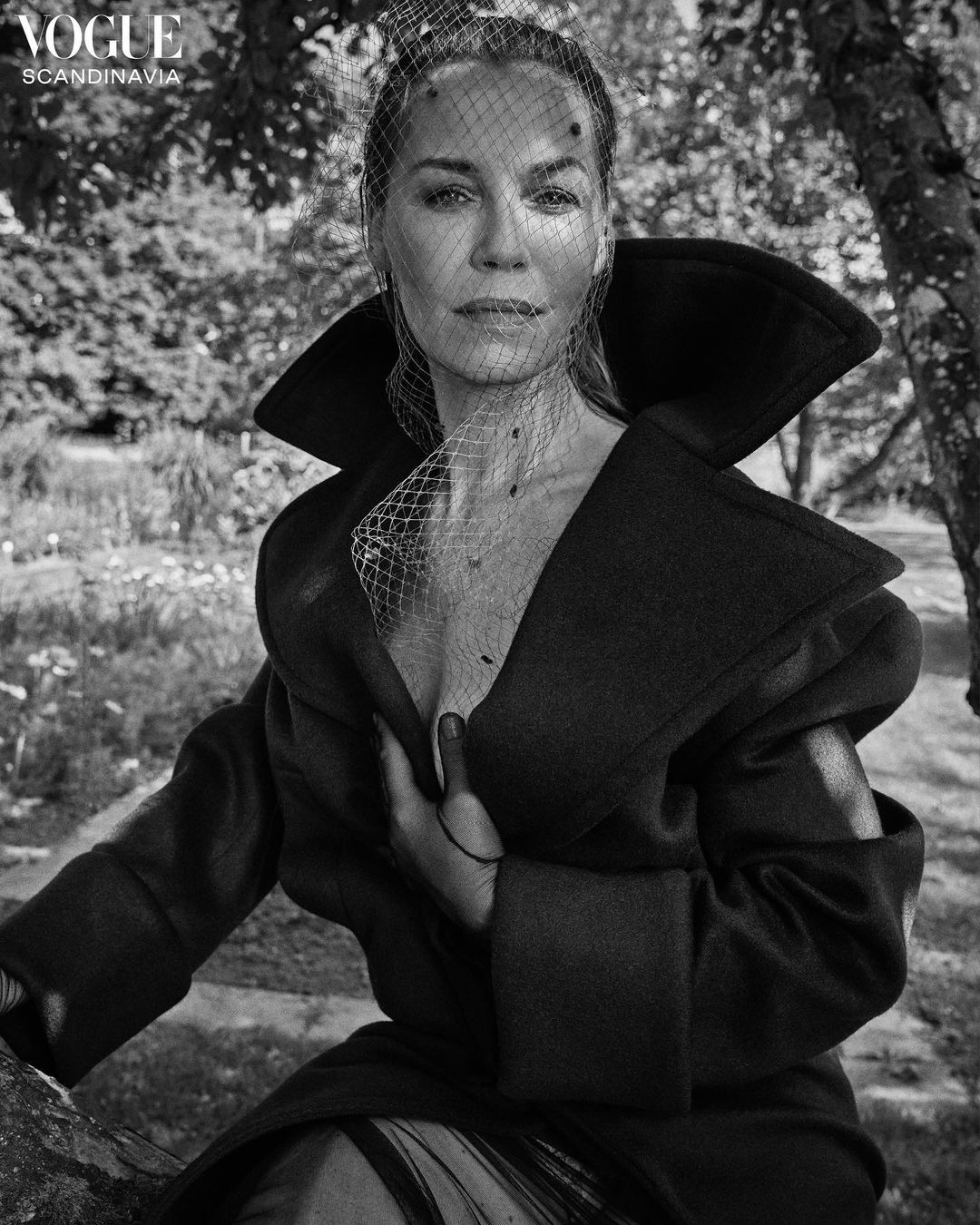 Monochrome portrait of a woman in a dark coat and veil, set against a blurred outdoor backdrop. Vogue Scandinavia logo visible.