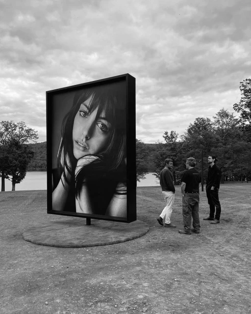 Large outdoor portrait of a woman displayed near group of men, with a cloudy sky and trees in the background, captured in black and white.