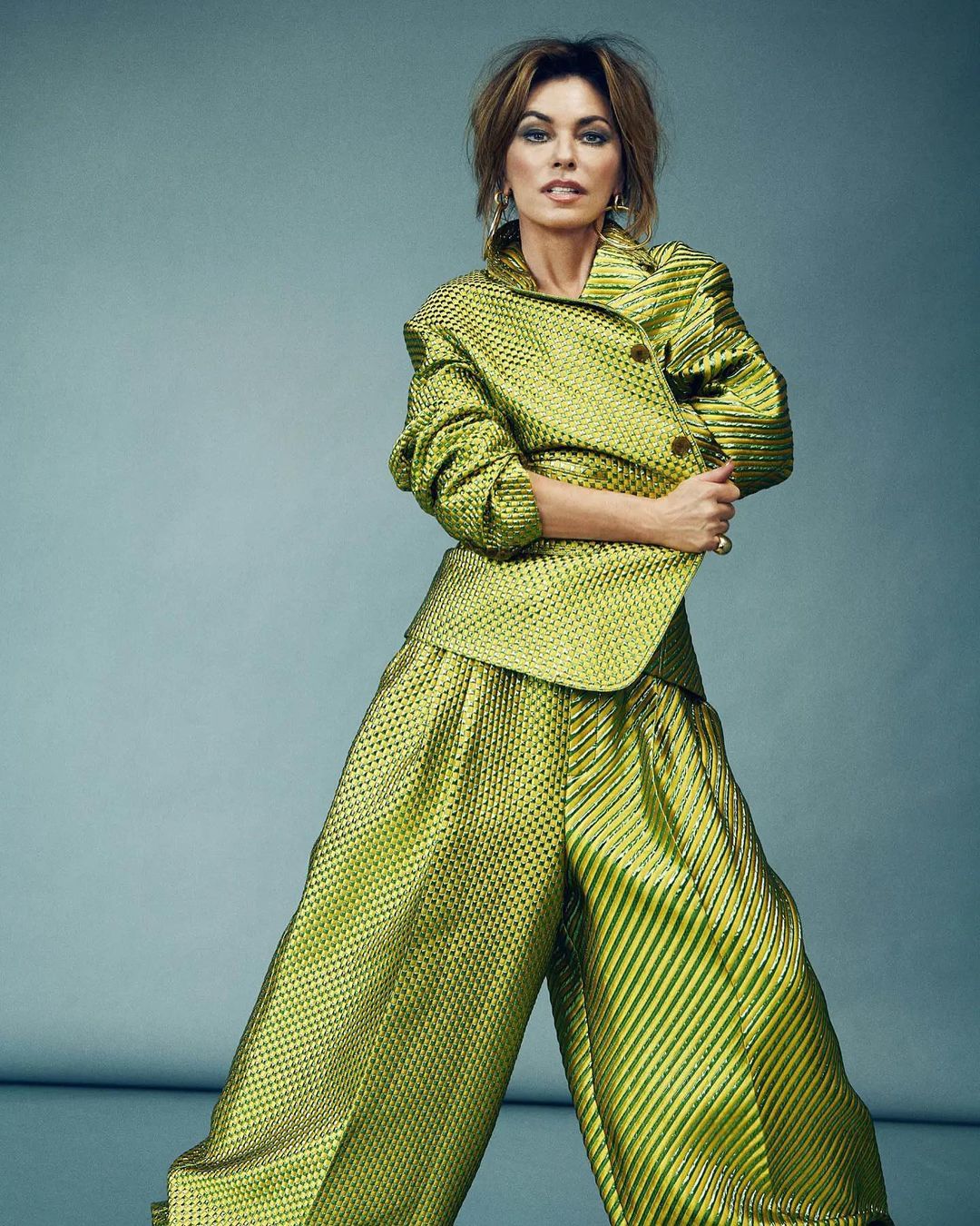Shania Twain in a green metallic suit posing in front of a muted blue background. The singer wears a jacket and wide leg pants.