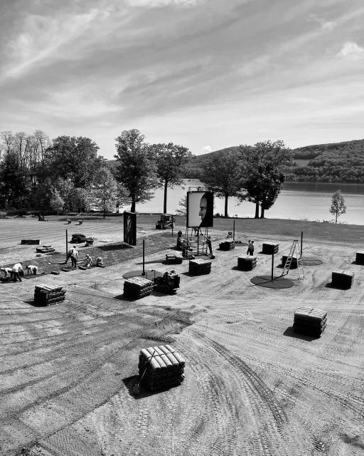 Workers install large portraits at an outdoor art installation near a lake, featuring black and white photography and stacked hay bales.