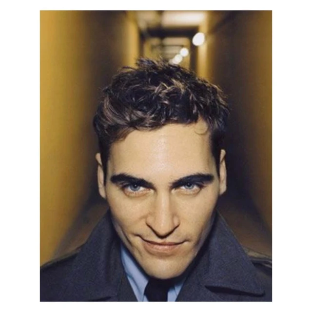 Close-up studio portrait of a man with dark wavy hair and blue eyes wearing a coat and shirt, illuminated by warm lighting against a dark hallway.