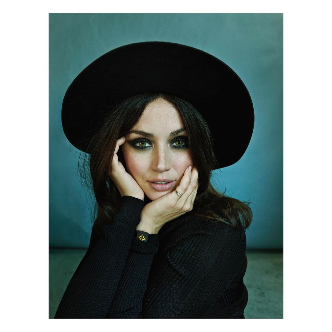 Portrait of Ana de Armas wearing a wide-brimmed black hat and dark ribbed turtleneck, framed against a textured blue backdrop.