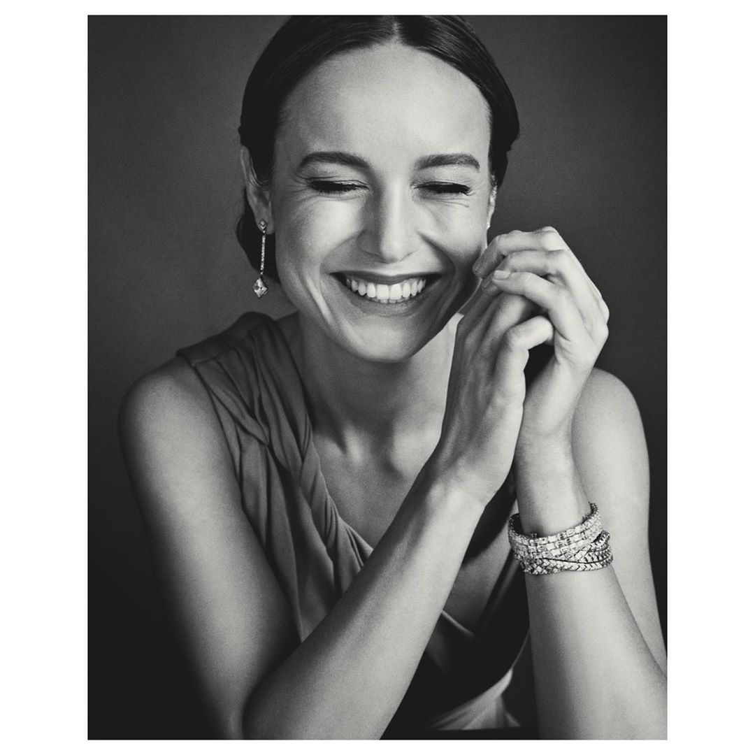 Black and white portrait of a smiling woman with dark hair wearing jewelry. She is indoors against a dark background.