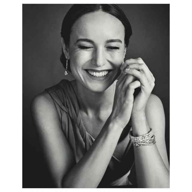 Black and white portrait of a smiling woman with dark hair wearing jewelry. She is indoors against a dark background.