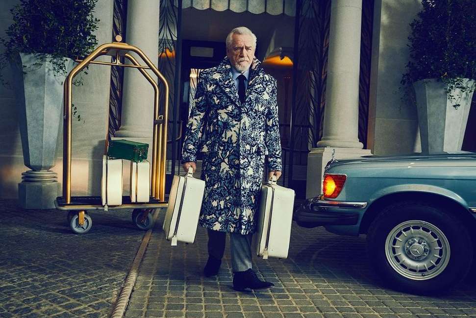 Mature man in patterned coat carries matching suitcases in front of luxury hotel, standing near bellhop luggage cart and vintage car.