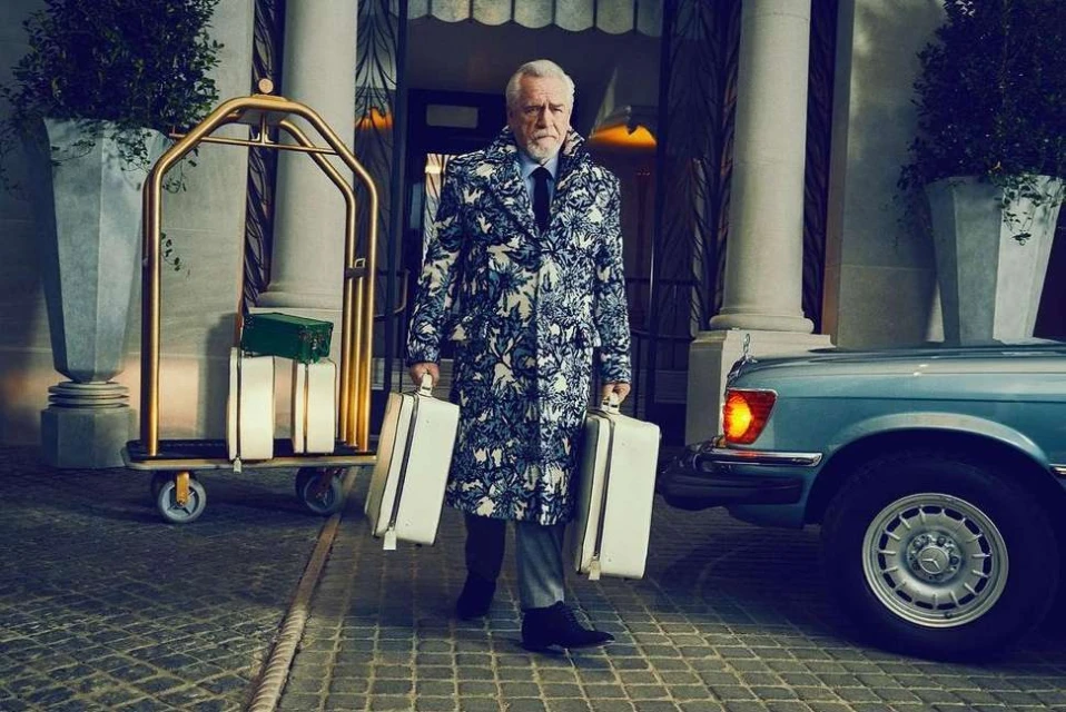 Mature man in patterned coat carries matching suitcases in front of luxury hotel, standing near bellhop luggage cart and vintage car.