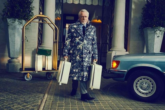 Mature man in patterned coat carries matching suitcases in front of luxury hotel, standing near bellhop luggage cart and vintage car.