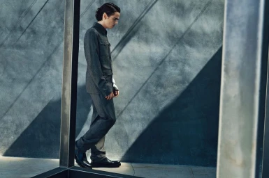 Man in a grey suit jacket and trousers walking past a textured concrete wall with angled shadow lines.