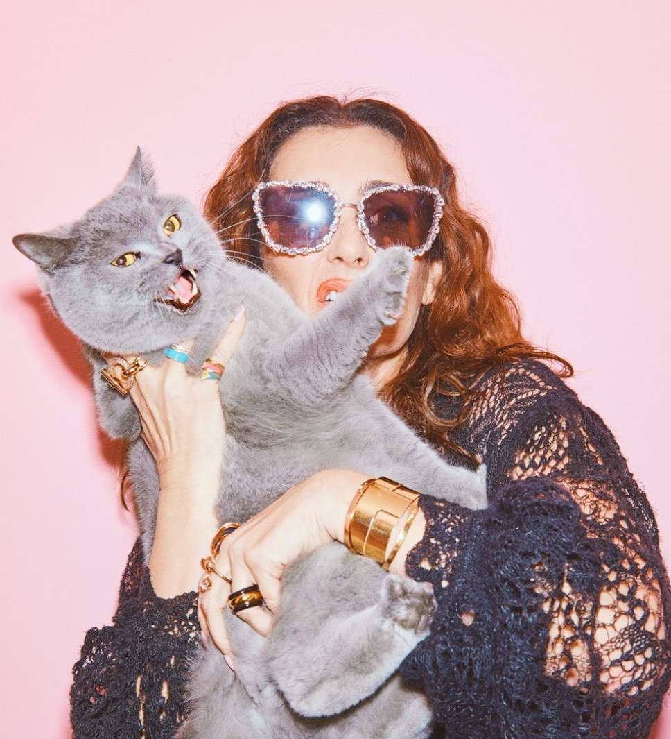 An adult female holding a gray cat with mouth open in a studio against a pink background, wearing sunglasses and a black crocheted top.