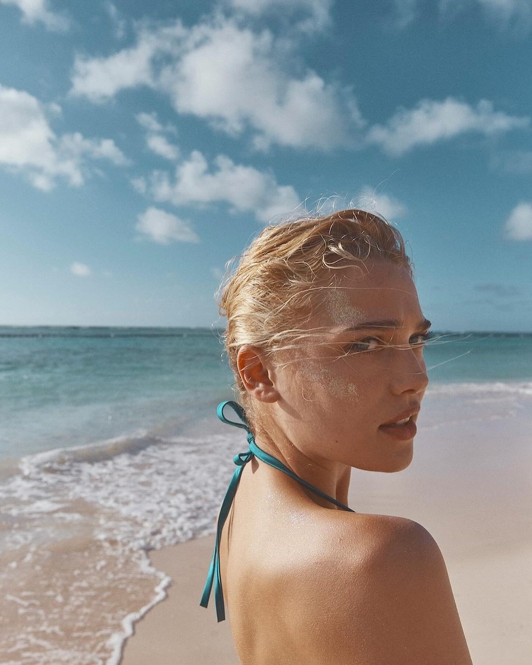 Woman with blonde hair wearing a halter top at the beach, posing with ocean and sky in the background, sunny day.