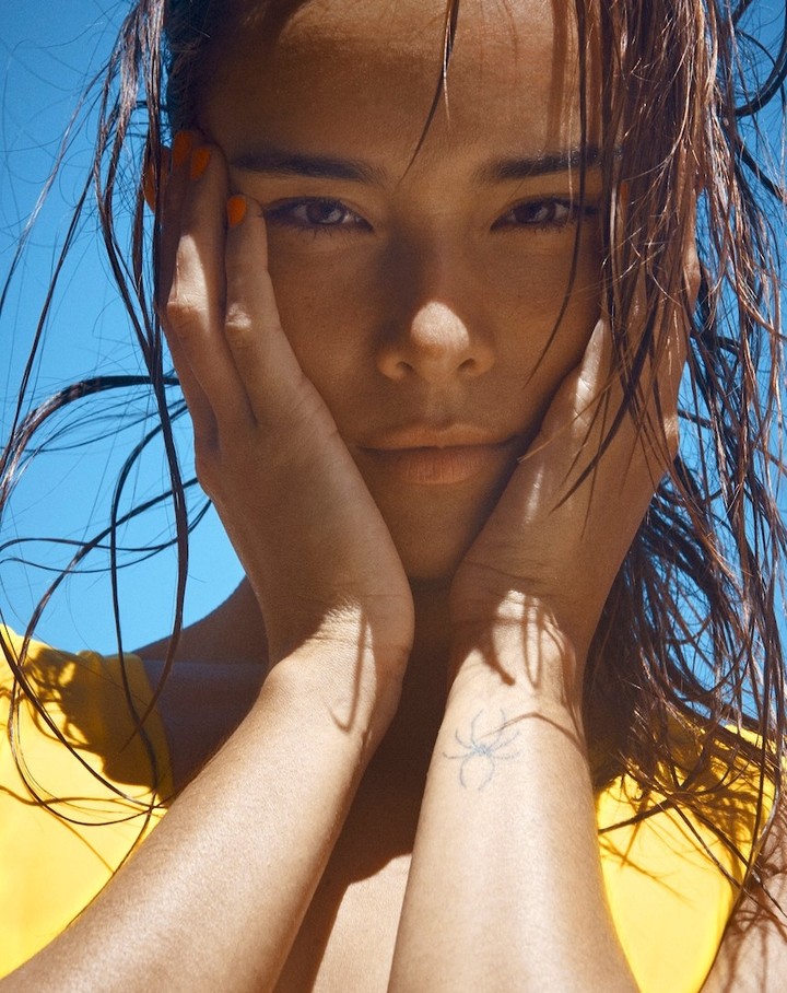 Close-up portrait of a woman with wet hair, tanned skin, and an arm tattoo against a vibrant blue sky, showcasing a fresh beauty aesthetic.