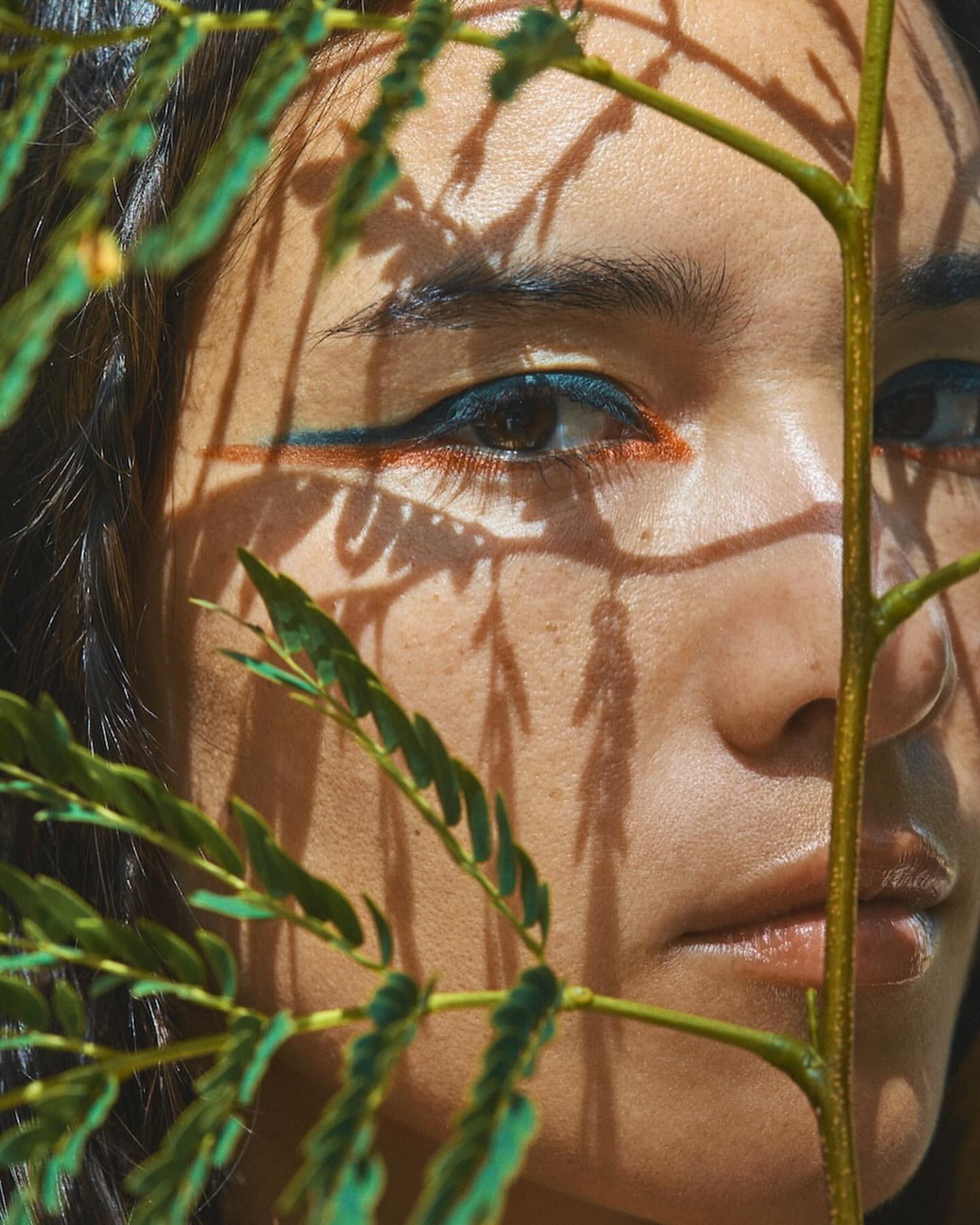 Close-up of a woman's face, partially obscured by a leafy branch, featuring vibrant blue and orange eyeliner, set in a sunlit outdoor scene.