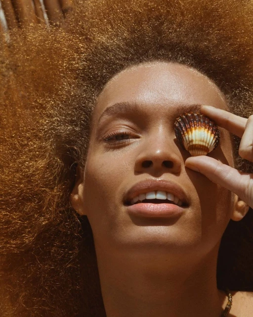 Portrait of a woman with coppery afro hairstyle lying in the sun, holding a small seashell over her eye in a playful, sunny scene.