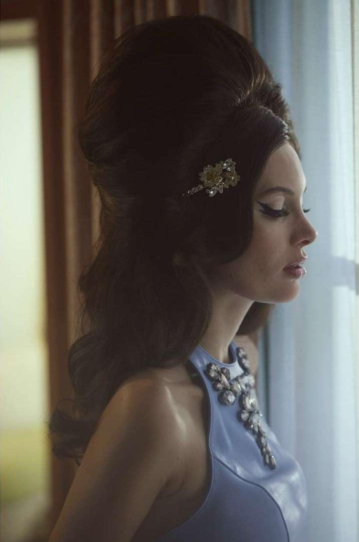 Glamorous woman with a large beehive hairstyle looks out a window wearing a jeweled blue dress in soft, diffused lighting.