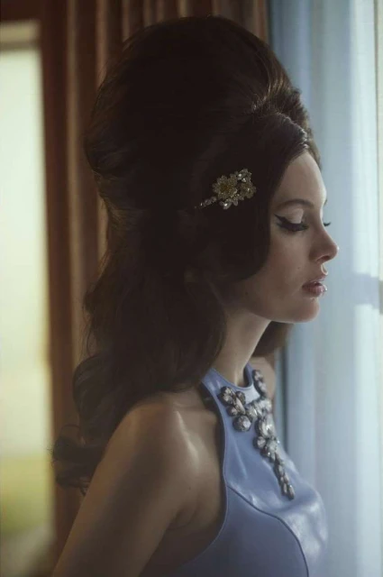 Glamorous woman with a large beehive hairstyle looks out a window wearing a jeweled blue dress in soft, diffused lighting.