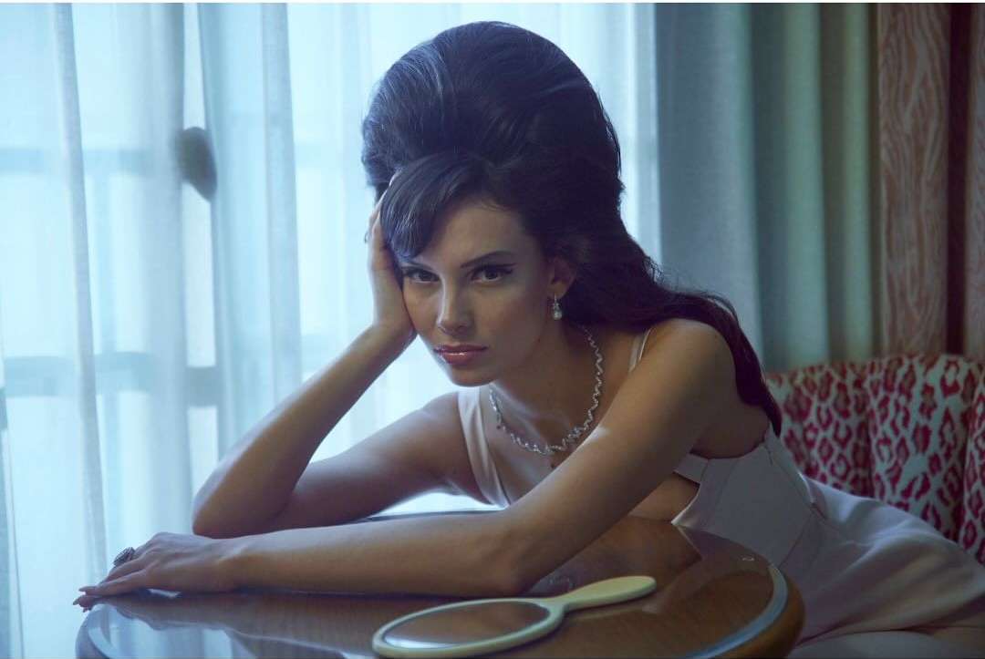 Portrait of a brunette woman with vintage-inspired high hair and jewelry, resting her head on her hand at a reflective table.