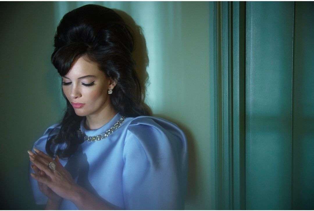 Elegant woman with an updo hairstyle and blue dress poses indoors against a blue-green wall, showcasing fashion and beauty.