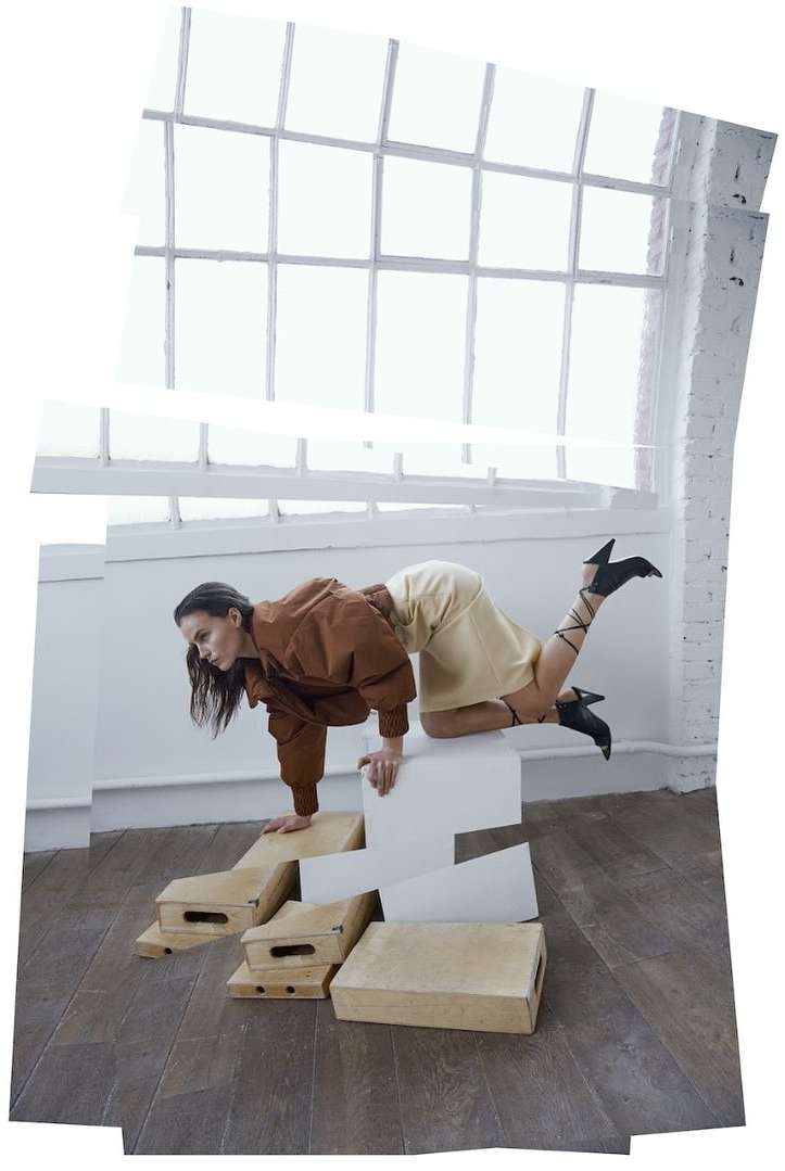 A woman wearing a brown jacket and light yellow skirt poses on white steps in front of a large window in a staged fashion shot.
