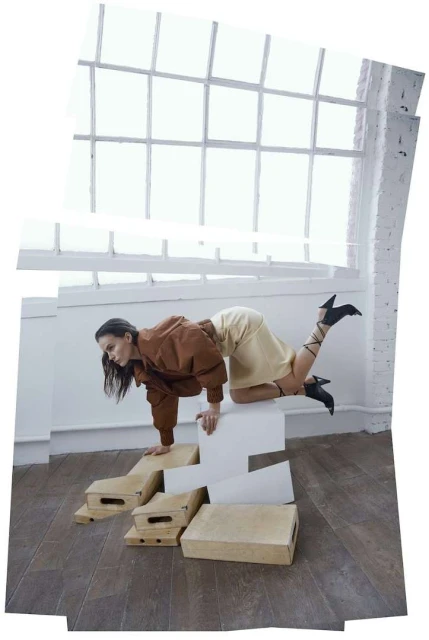 A woman wearing a brown jacket and light yellow skirt poses on white steps in front of a large window in a staged fashion shot.