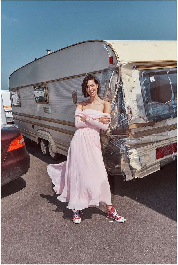 A young adult woman poses in a pink dress and red sneakers next to a weathered camper in an outdoor setting under a bright blue sky.