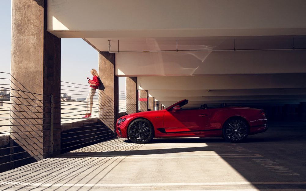 Red convertible parked in garage, with a woman in red leaning against railing looking at her phone near city buildings.