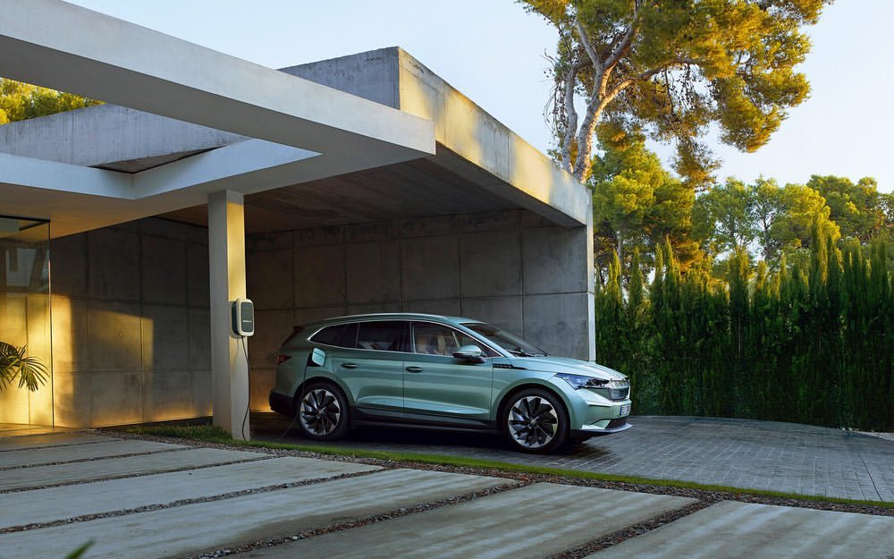 Green SUV parked and charging at a modern house with concrete driveway and lush green trees in the background during daytime.