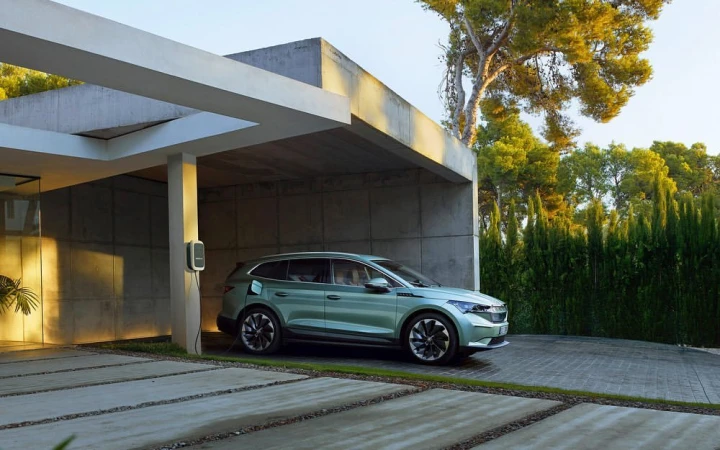 Green SUV parked and charging at a modern house with concrete driveway and lush green trees in the background during daytime.