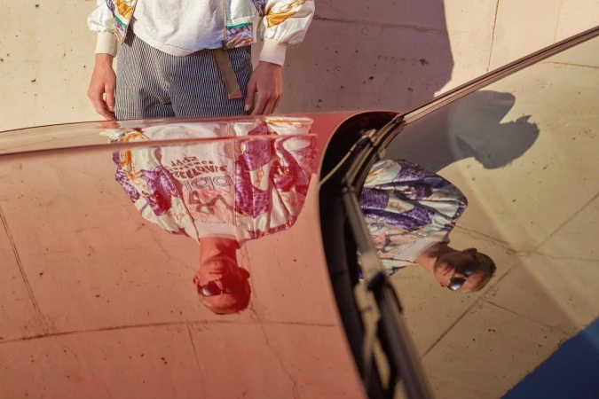 Reflection of a person wearing sunglasses and a patterned jacket on the red hood of a car under bright sunlight.