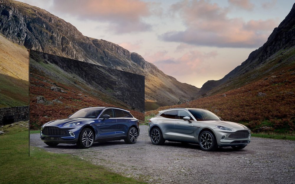 Two Aston Martin DBX707 SUVs in silver and blue parked on a gravel road with a mountain and cloudy sky background, automotive shoot.