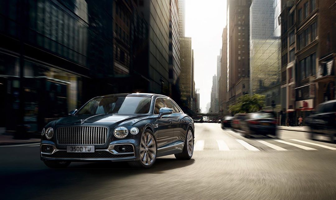 Luxury dark gray Bentley sedan driving on city street with motion blur, surrounded by tall buildings on a sunny day.