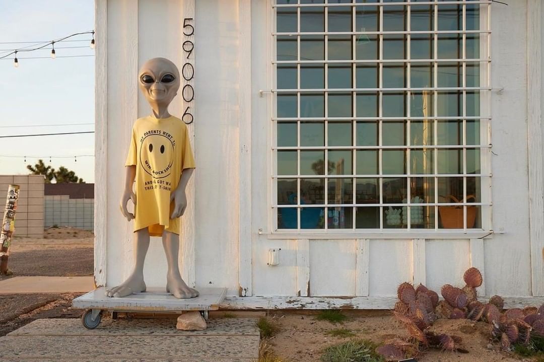 Grey alien mannequin wearing a yellow smiley face t-shirt stands outside a white building with a barred window on a sunny day.