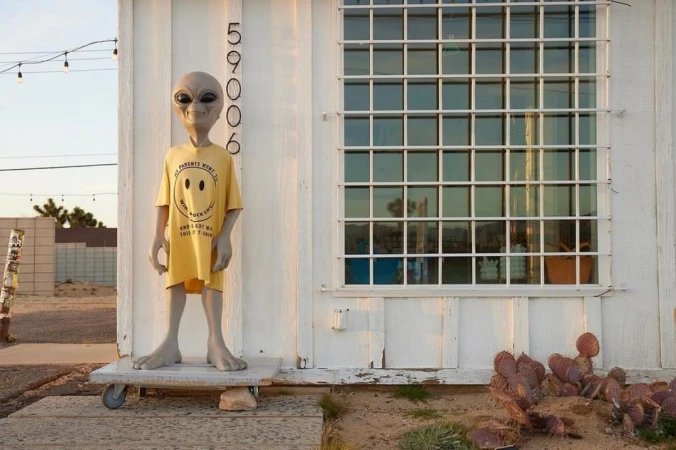 Grey alien mannequin wearing a yellow smiley face t-shirt stands outside a white building with a barred window on a sunny day.