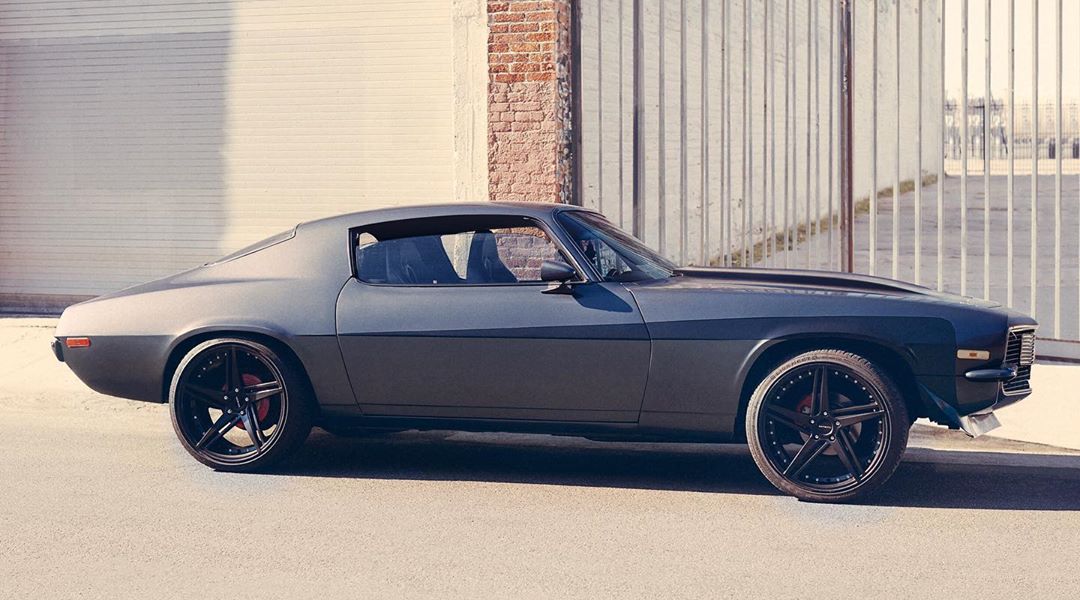 Side view of a matte gray classic car with black rims parked against a wall and metal gate, bathed in bright sunlight. Automotive.