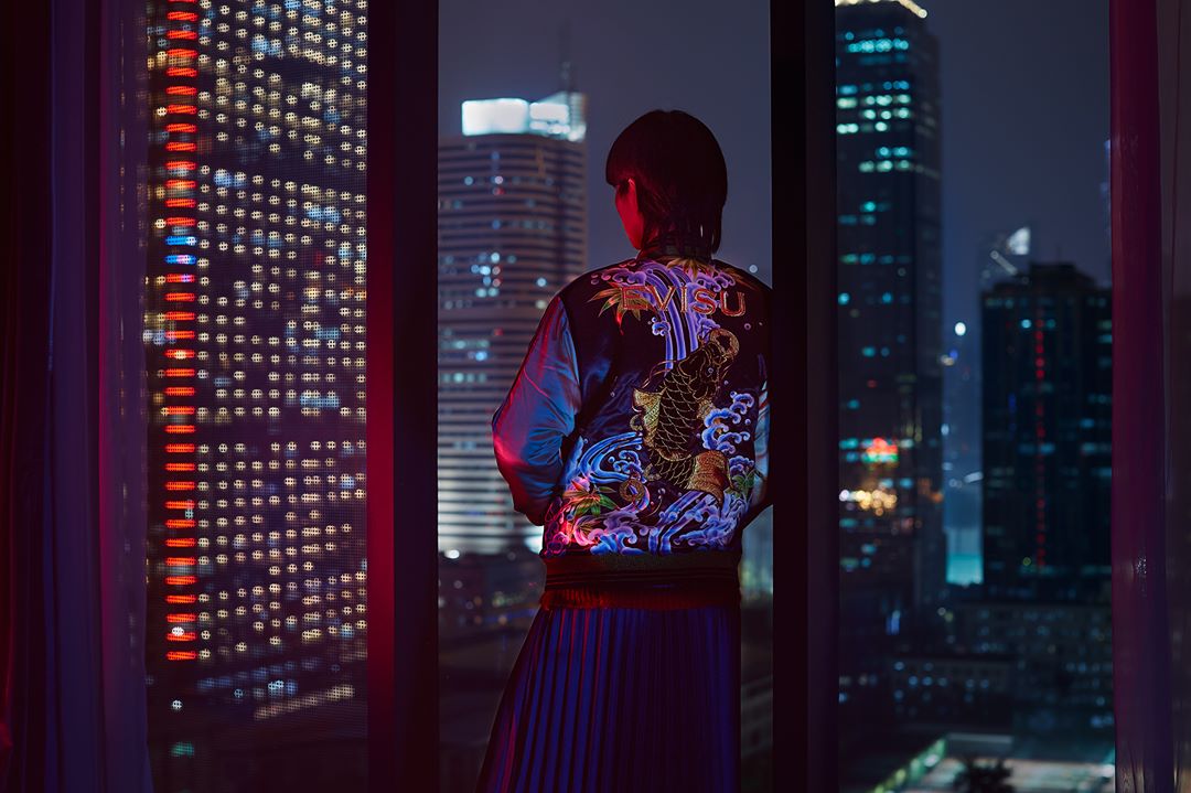 Adult viewed from behind, wearing a patterned jacket, gazing out a window at a sparkling cityscape during the dark of night.