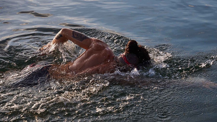 Adult male swimmer with a muscular build is swimming in blue water with arm raised, creating splashes; outdoor, daylight conditions.