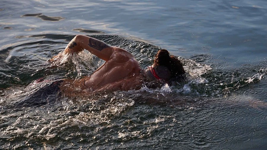 Adult male swimmer with a muscular build is swimming in blue water with arm raised, creating splashes; outdoor, daylight conditions.