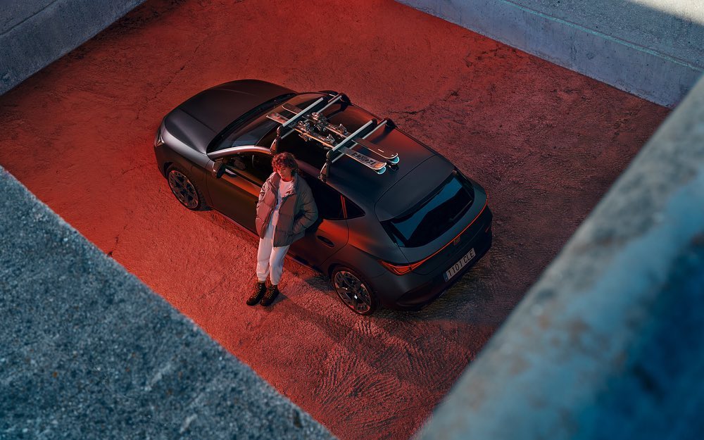 Person stands by a dark grey Cupra Formentor with skis on the roof rack in an outdoor urban setting with red and blue lighting.