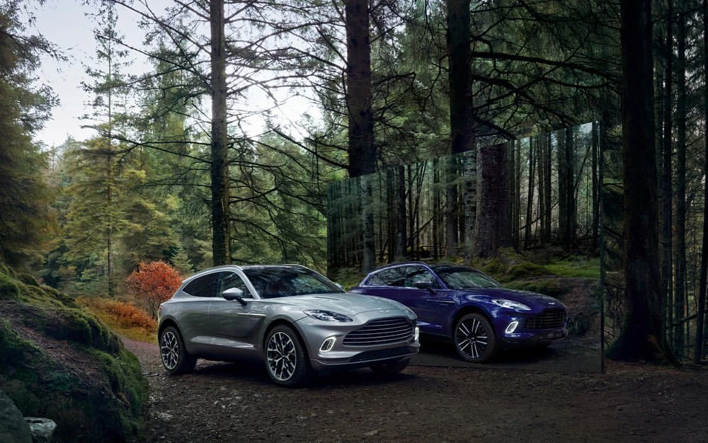 Two Aston Martin DBX707 cars are parked on a dirt path in a lush forest. One is silver, the other is blue. Tall trees surround the vehicles.