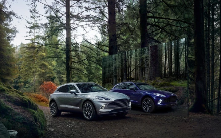 Two Aston Martin DBX707 cars are parked on a dirt path in a lush forest. One is silver, the other is blue. Tall trees surround the vehicles.