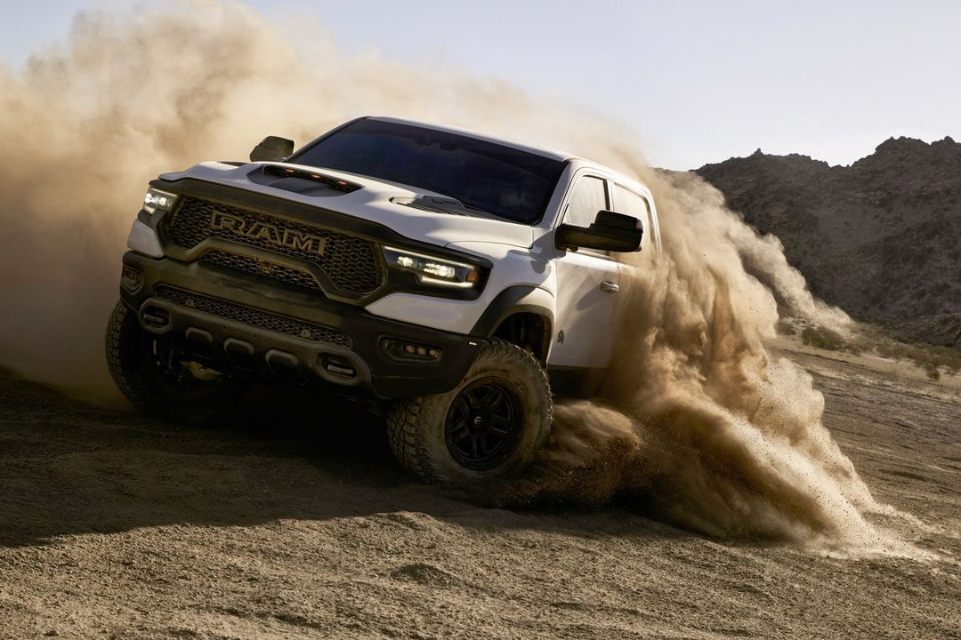 A white Ram TRX truck drives through a desert landscape, kicking up a cloud of dust while in motion, with mountains in background.