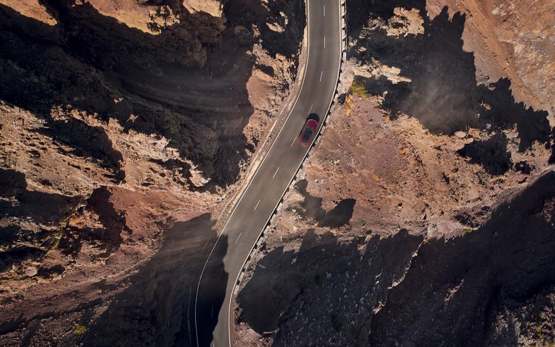 Aerial view of a red car driving along a winding asphalt road through a rocky canyon landscape, captured from a high angle.