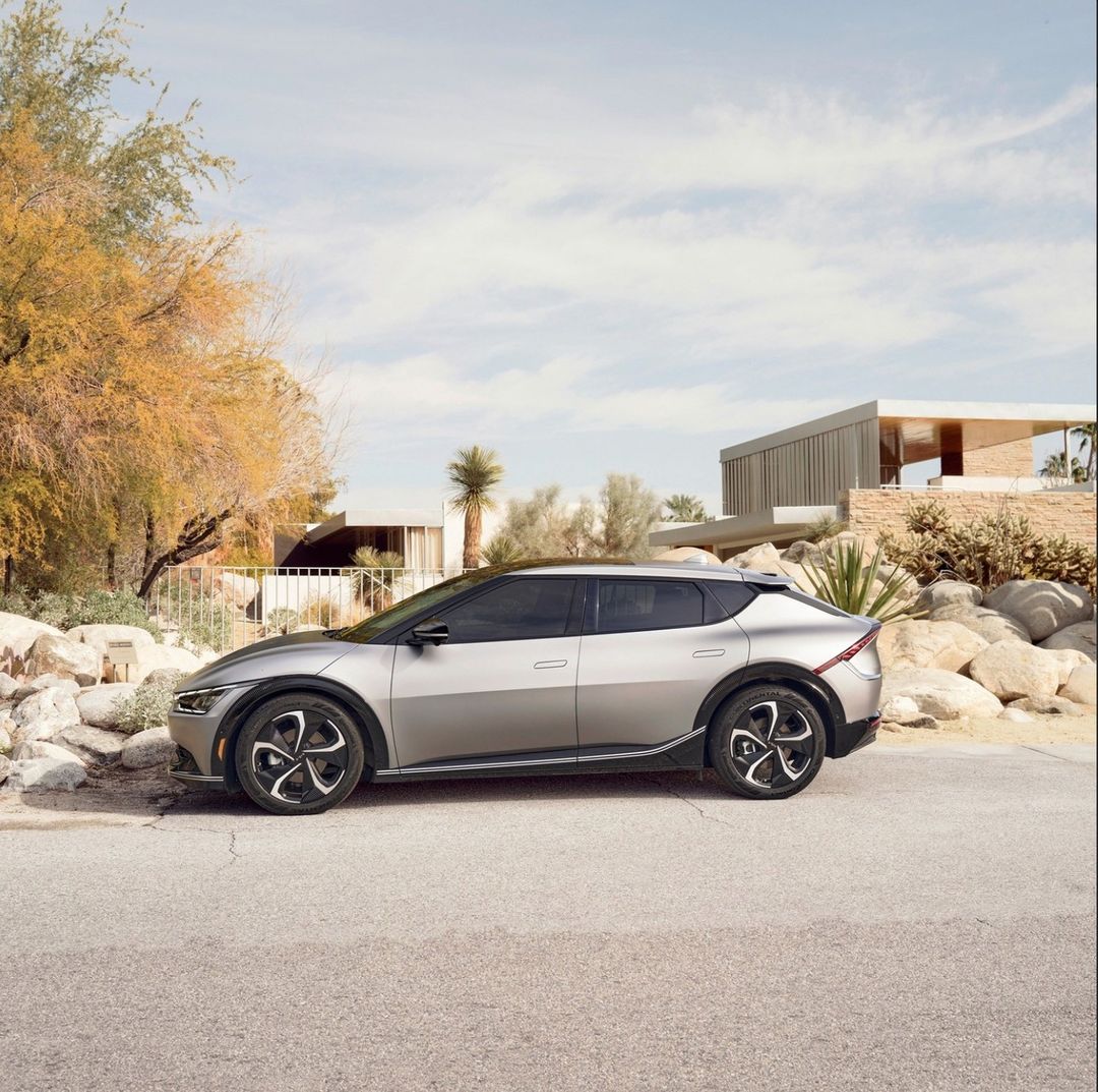 Silver Kia EV6 electric SUV parked on an asphalt road, against a backdrop of mid-century modern house and arid desert plants.