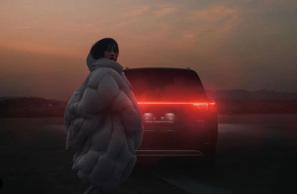 A person in a white puffer jacket stands next to a dark SUV against a dusky sky. Automotive style and outdoor fashion meet.