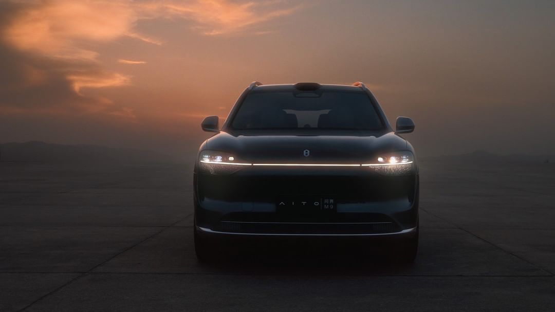 Front view of a dark AITO SUV with headlights illuminated against a dusky sky at sunset. The car is parked on a dark surface.