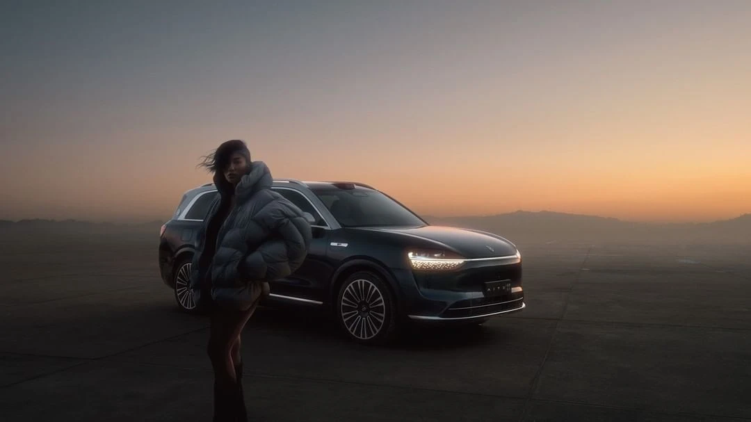 A woman stands in front of a dark blue SUV in a desert landscape at sunset. The woman wears a puffy coat and the car's headlights are on.